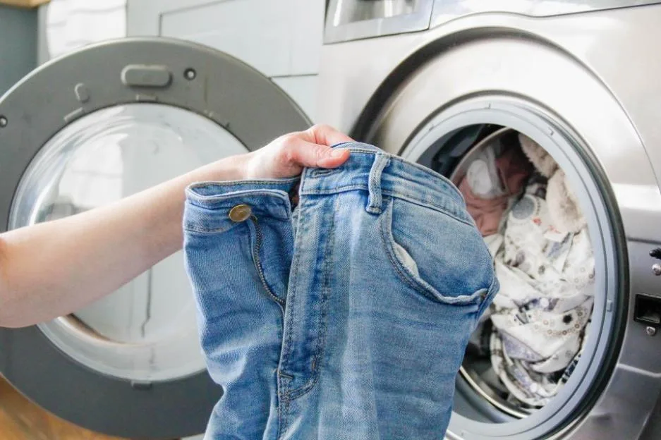 A person holding a pair of blue denim jeans in front of an open washing machine.