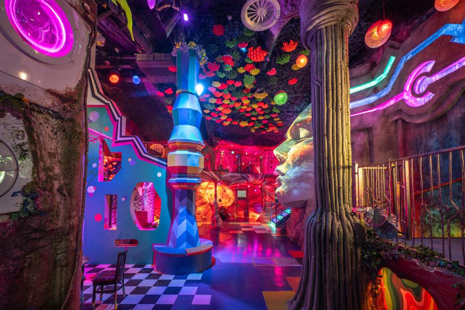 A brightly lit, psychedelic indoor art exhibit with neon colors, a giant face sculpture, and a towering structure covered in geometric patterns.
