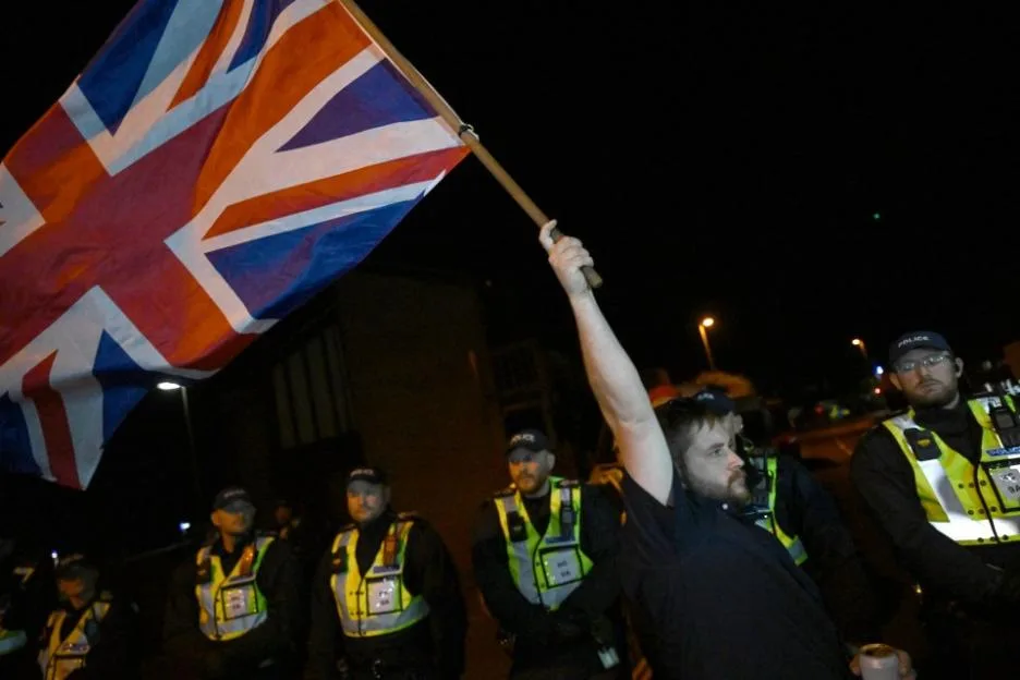 A protestor holds a Union Jack flag during an anti-migrant protest in Gosport, England, surrounded by police officers at night.