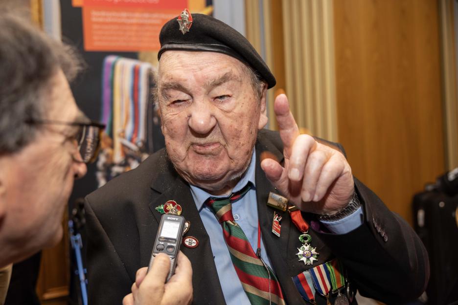 Richard Aldred, a 99-year-old World War II veteran, wearing a beret and military medals, speaks to a reporter holding a Sony voice recorder.