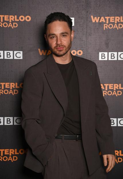 Actor Adam Thomas, in a dark brown suit, poses in front of a "Waterloo Road" backdrop.