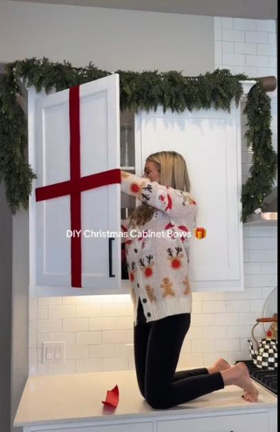 Woman decorating a kitchen cabinet door with red ribbon to resemble a Christmas present.