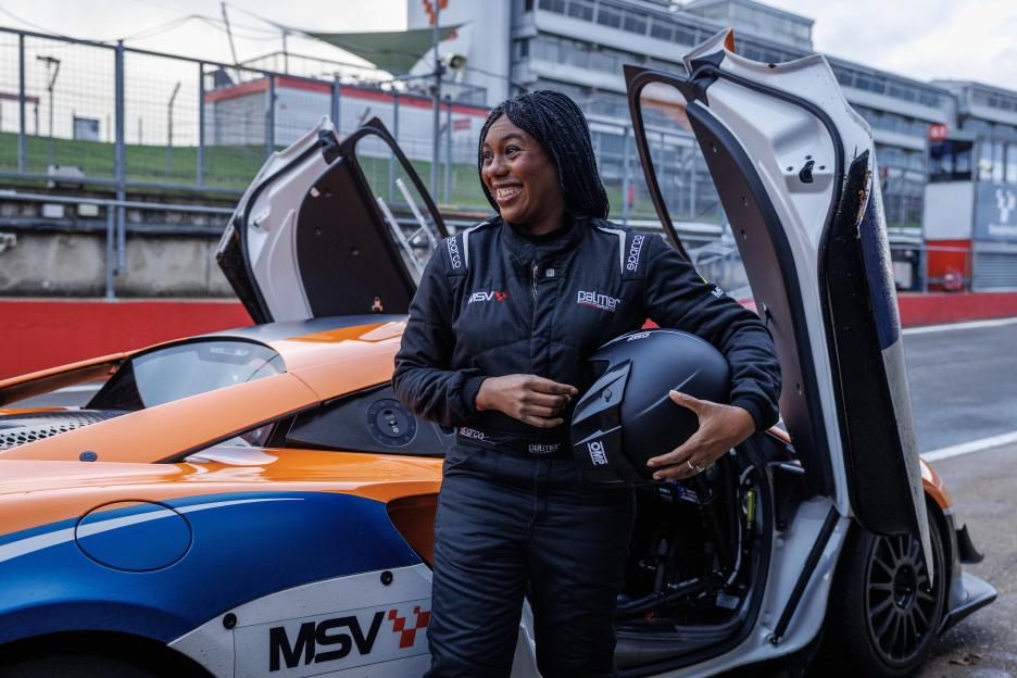 Kemi Badenoch, dressed in a racing suit and holding a helmet, stands next to a McLaren race car.