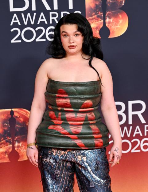 Lola Young attends The BRIT Awards 2026, wearing a dark green strapless top with red and black patterned cutouts, and patterned pants with small padlock adornments.