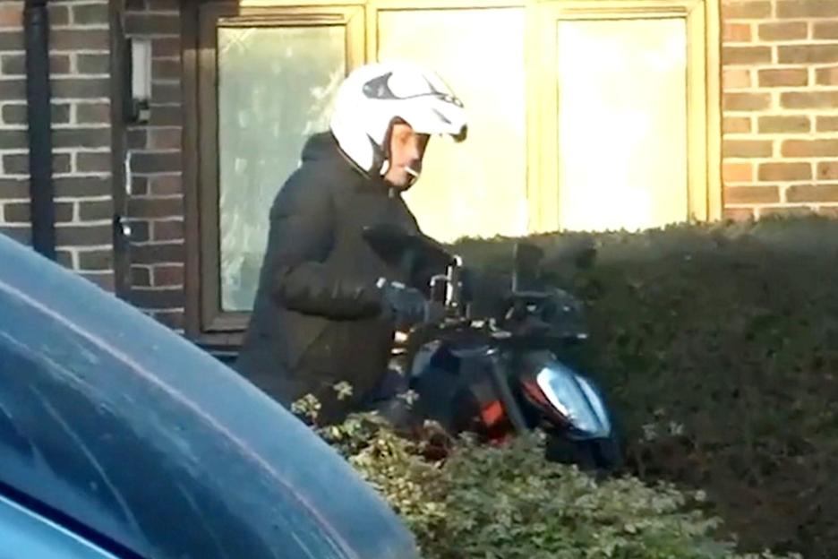 A man in a white helmet and dark jacket, with a cigarette in his mouth, sitting on a motorcycle partially hidden by bushes, in front of a brick building.