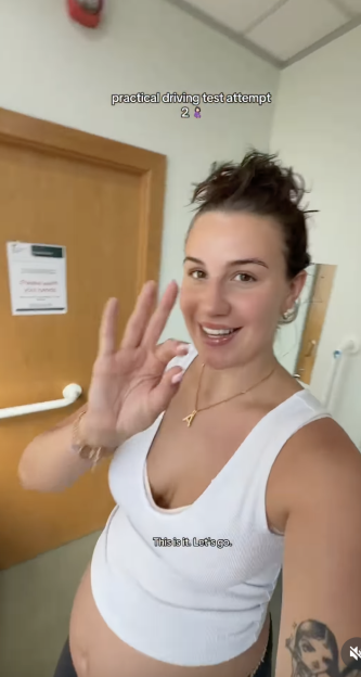 A pregnant woman smiling and holding up an "OK" hand sign before her second driving test attempt.
