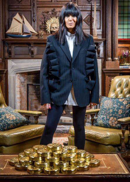 A woman in a black jacket and leggings stands behind a pile of gold coins on a table.