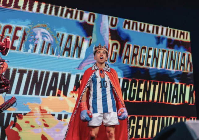 Boxer with a crown and red cape over an Argentinian jersey, standing in front of a screen displaying "ARGENTINIAN KING".
