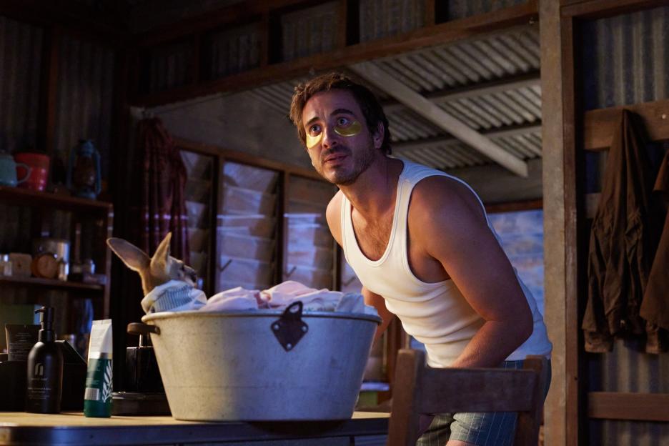 A man wearing golden eye masks and a white tank top looks at a baby kangaroo in a bucket.