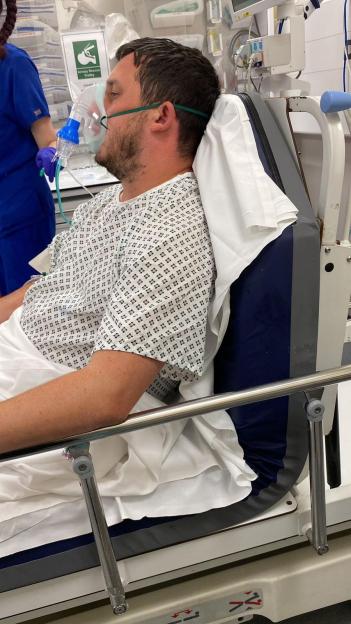 Man in a hospital gown and oxygen mask, sitting in a hospital bed.