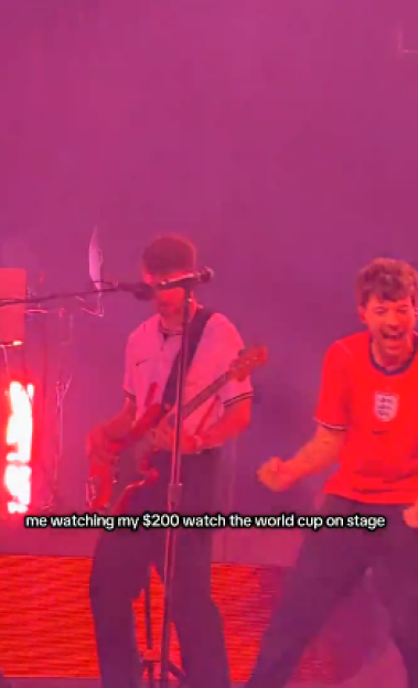 Two musicians on stage under red lights, one playing guitar, the other singing and wearing an England jersey.