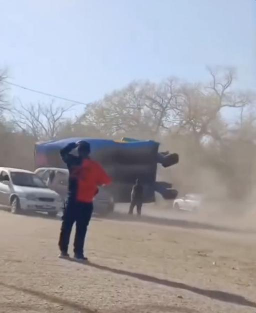 A man watching as a bouncy castle is sent flying by strong winds, creating a dust cloud on a dirt road.