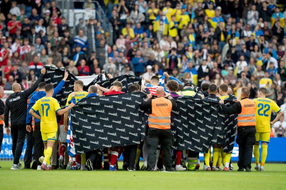 A group of football players huddled together on the field under a black blanket with "Albani" written on it.
