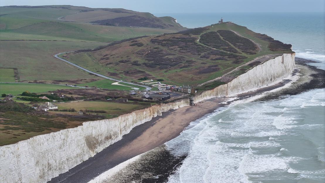 Aerial view of the white cliffs of Birling Gap with a narrow sandy beach and foamy ocean waves, with green hills and a lighthouse in the background.