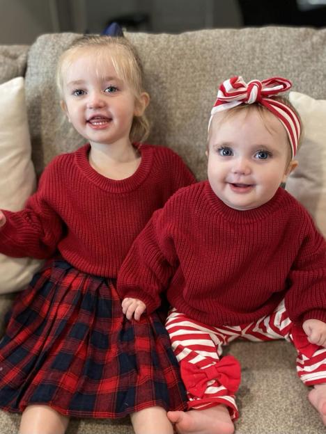 Two young girls, both wearing red tops, sit on a couch.