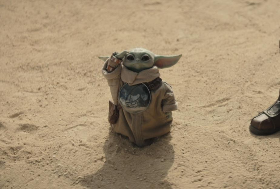Grogu standing on sand next to The Mandalorian's leg.