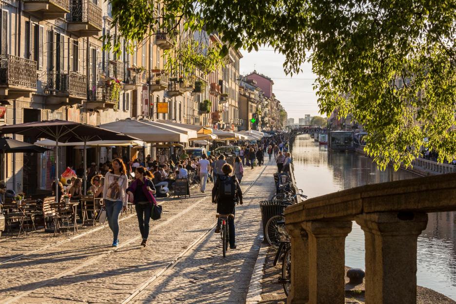 The Naviglio Grande canal in Milan, with people walking and dining at outdoor cafes along the cobbled path, Alzaia Naviglio Pavese.