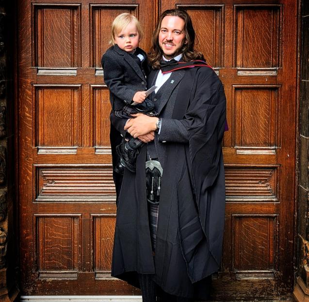 A man in a graduation gown and kilt holds a young boy in a suit and kilt.