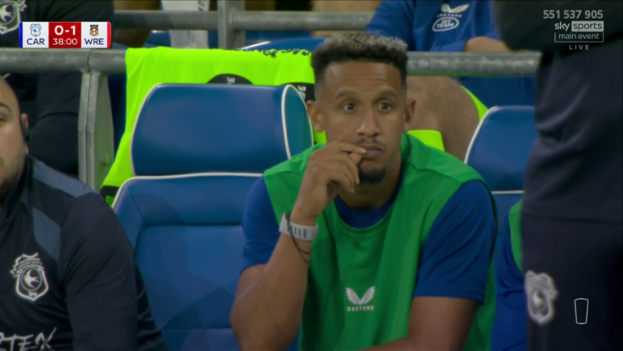 A male soccer player in a blue shirt and green vest sits on the bench looking concerned, with a game scoreboard in the upper left showing "CAR 0-1 WRE" at 38:00.