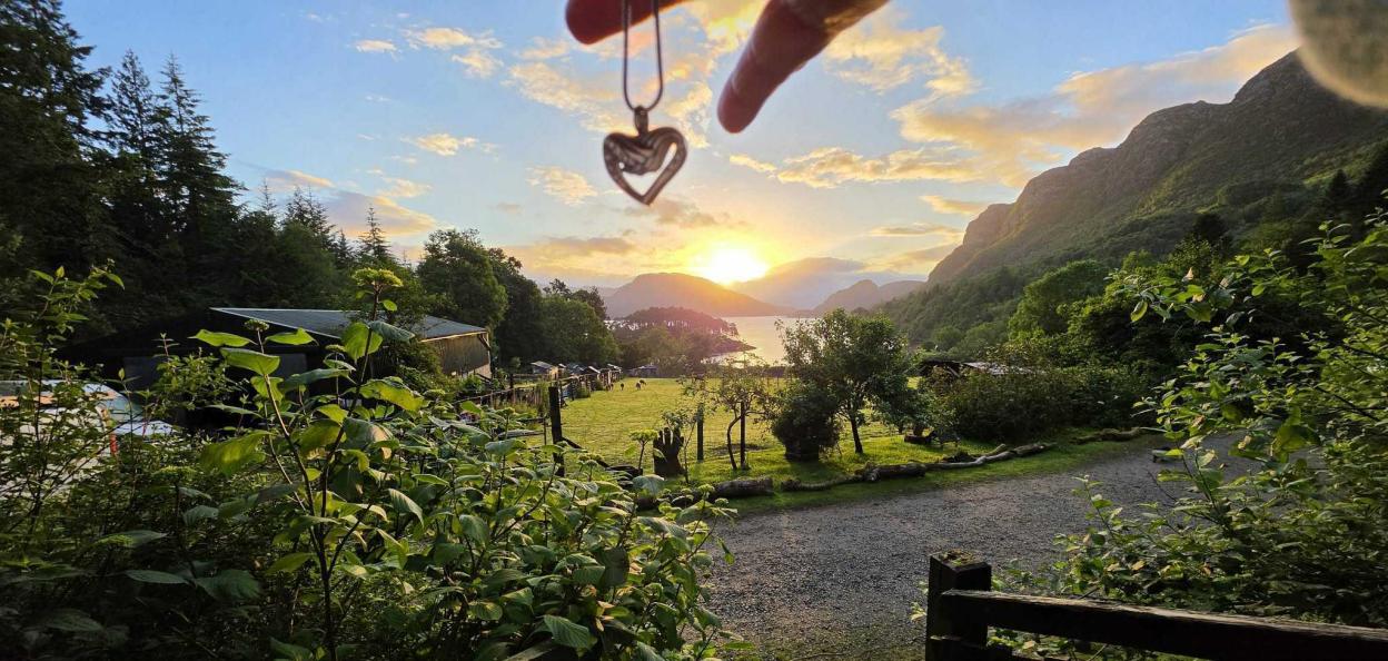 A hand holding a heart-shaped pendant in front of a sunset over a body of water surrounded by mountains and trees.