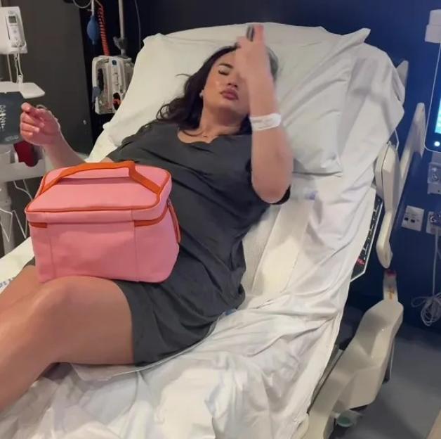 A woman wearing a dark grey dress and makeup lying in a hospital bed with a pink lunchbox on her lap.