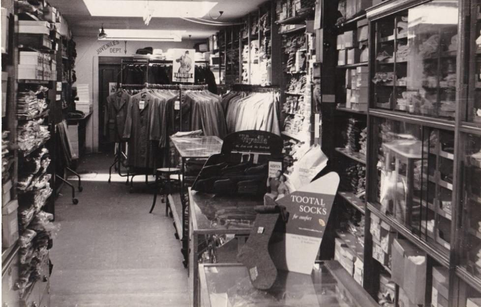 Interior of a small haberdashery or men's clothing shop in black and white, with racks of coats and shelves of boxed items and a sign for "Tootal Socks" in the foreground.