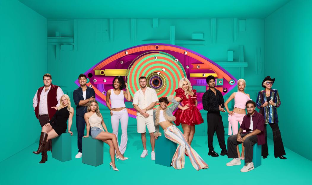 A group of fourteen Big Brother contestants pose in front of a giant eye logo.
