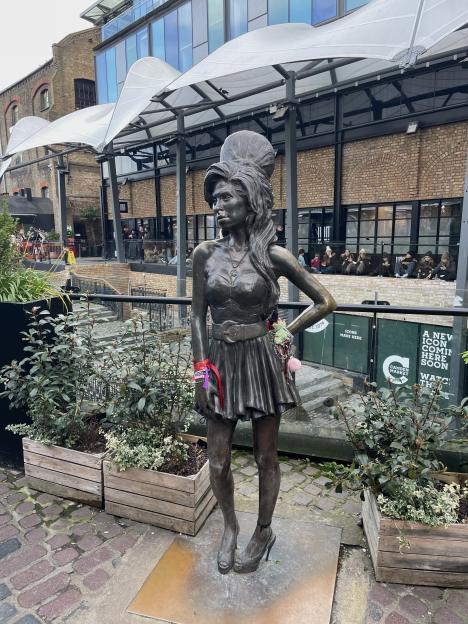 A bronze statue of Amy Winehouse wearing a short dress and high heels stands outdoors, with colorful bracelets and ribbons tied to her wrists.