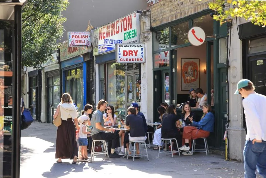 People sitting at outdoor tables on a sunny London street, outside shops including Newington Green Dry Cleaners and a Polish Deli.