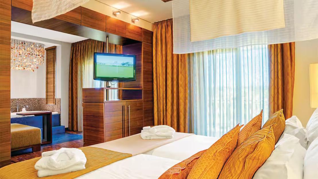 A hotel room with a TV, a bed with white sheets and golden pillows, and a view of an outside area through large windows.