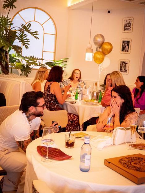A man proposes to a woman at a restaurant, surrounded by friends.