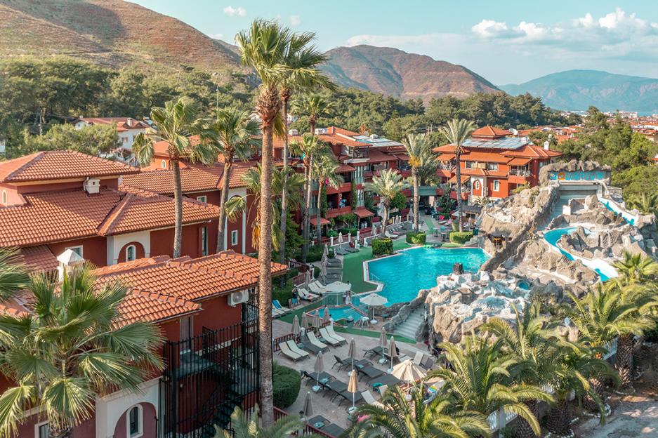 Aerial view of the Grand Aquarium Hotel in Dalaman, Turkey, with red-roofed buildings, swimming pools, waterslides, palm trees, and mountains in the background.