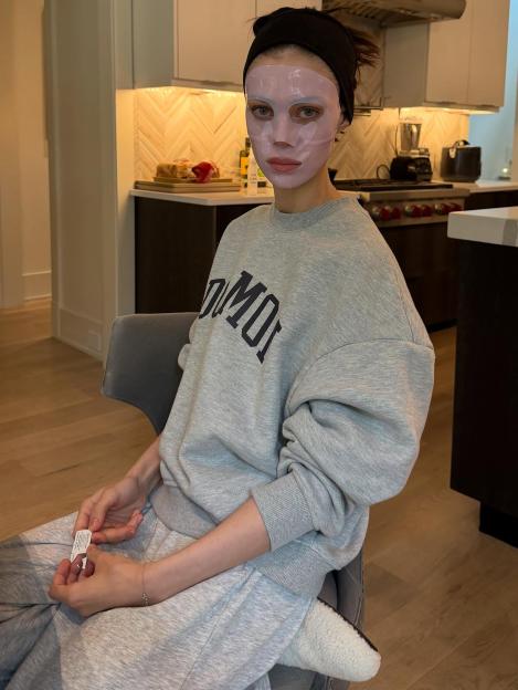 Nicola Peltz Beckham wearing a pink face mask and grey loungewear, sitting in a kitchen.