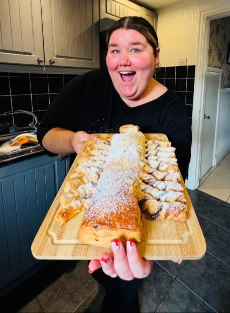 Becki Jones holds up a large, sugar-dusted pastry on a wooden board.