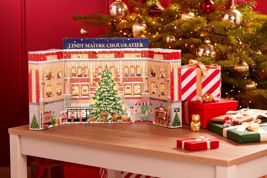 A Lindt Advent Calendar, shaped like a decorated building with a Christmas tree in front, next to wrapped gifts and chocolate treats on a table, with a Christmas tree in the background.
