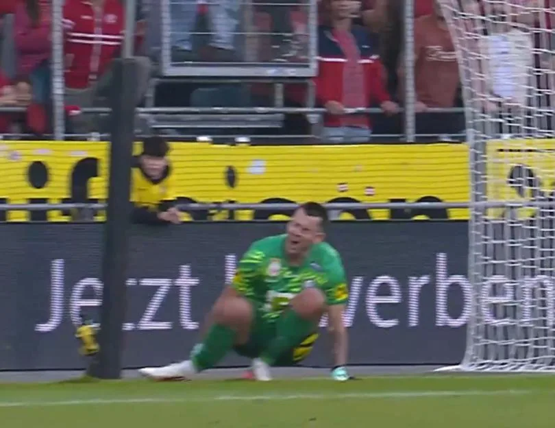 A goalkeeper in a green uniform sits on the pitch in agony, holding his leg near the goal.