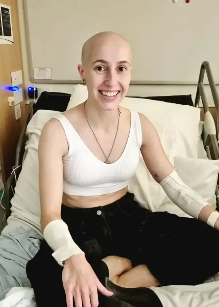 A young woman with a shaved head smiles while sitting up in a hospital bed, wearing arm splints.
