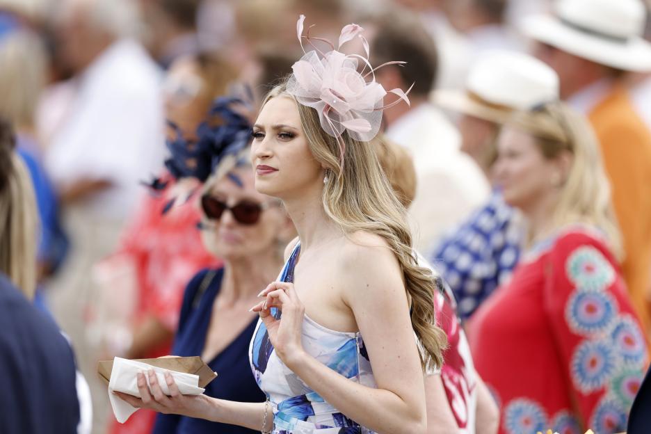 A racegoer with a pink fascinator, blue and white dress, and a food tray in hand, looking to her left at the Qatar Goodwood Festival.