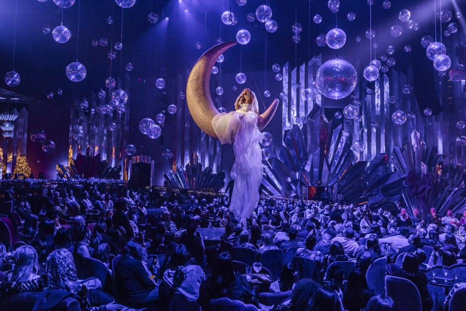 Leona Lewis performing in a purple-lit venue, singing while seated on a crescent moon prop, surrounded by hanging orb decorations, with an audience watching.