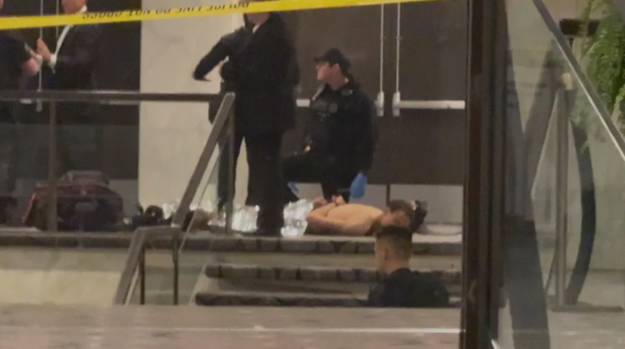 A man with a bare torso is lying face down on a concrete stair landing, handcuffed with a police officer kneeling nearby, another standing over him, and a yellow police tape above.