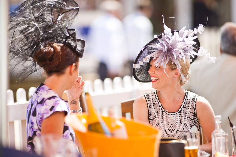 Two women in elaborate hats are sitting and talking at an outdoor event.