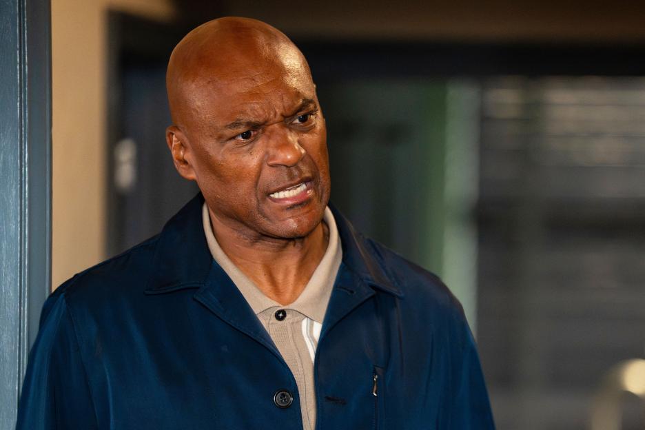 A bald man in a blue jacket looks angry with furrowed brows.