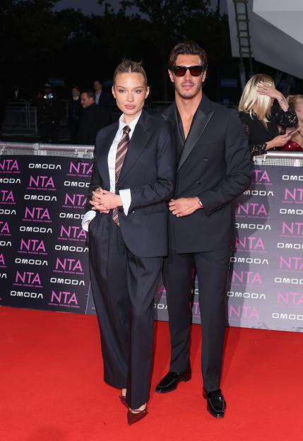 Julia Mayska in a dark pinstripe suit and tie next to Lorenzo Alessi in a dark tuxedo and sunglasses on a red carpet.