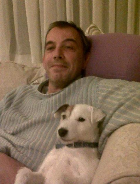 Keith Bridger sitting on a sofa with a small white dog with brown markings.
