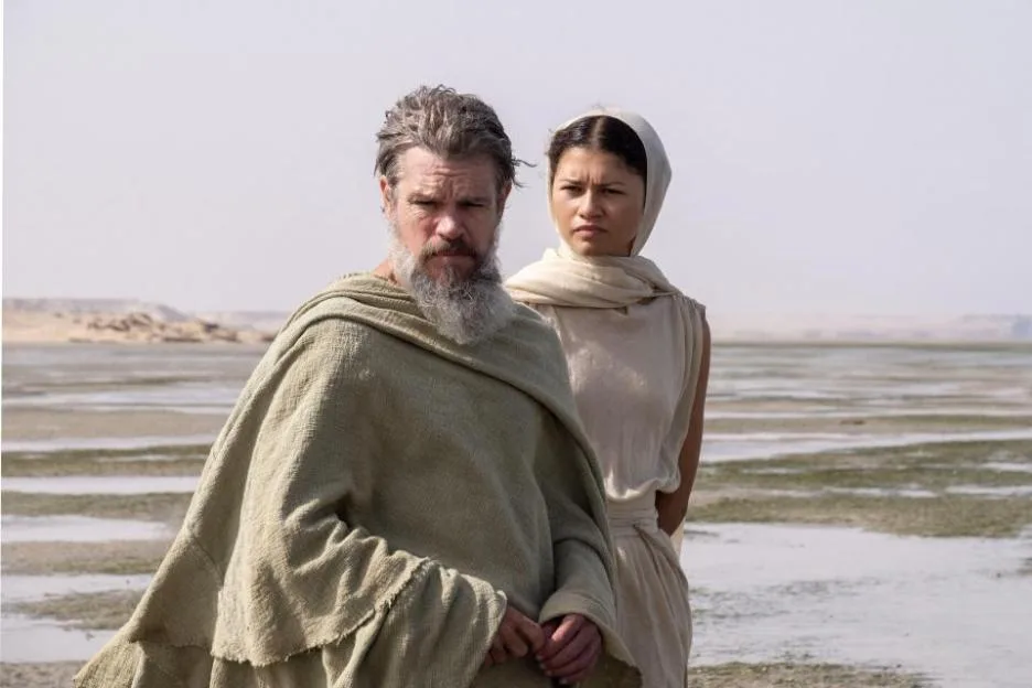 Matt Damon and Zendaya stand in a barren landscape, dressed in ancient-style draped garments.