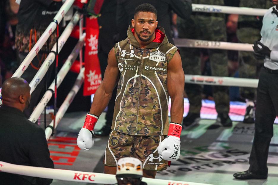 Boxer Anthony Joshua stands in a boxing ring, wearing a camouflage vest and shorts.