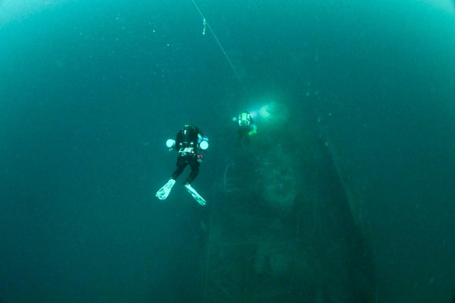 Two divers explore the shipwreck of the SS Nantes, with one diver's flashlight illuminating part of the vessel.