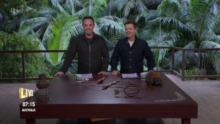 Ant and Dec standing behind a large wooden table with jungle foliage in the background.