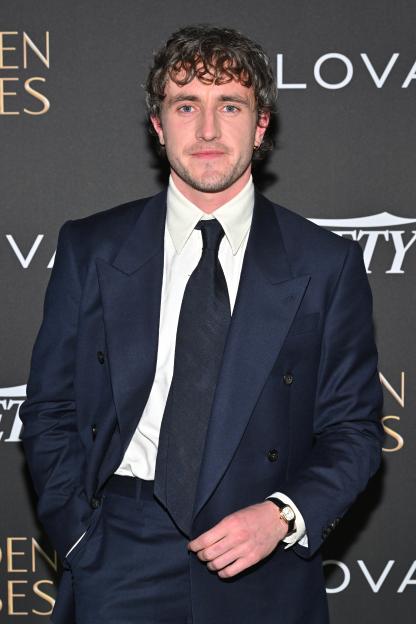 Paul Mescal in a navy suit and white shirt with a navy tie, standing in front of a black backdrop with white and gold lettering.