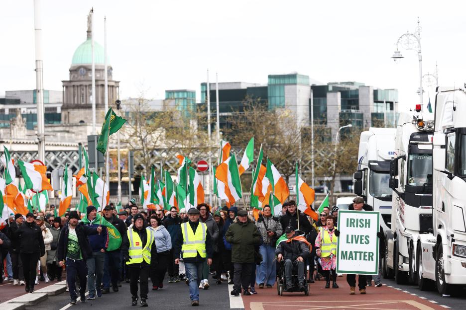 Protesters carrying Irish flags and a sign that reads "Irish Lives Matter" march in a street next to parked trucks.